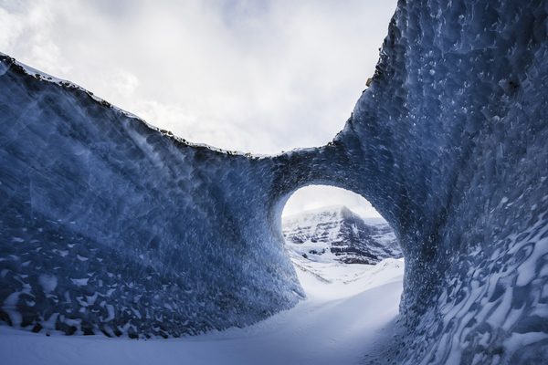 Round ice arch and a snow path leading out to a rocky peak under soft cloud.
